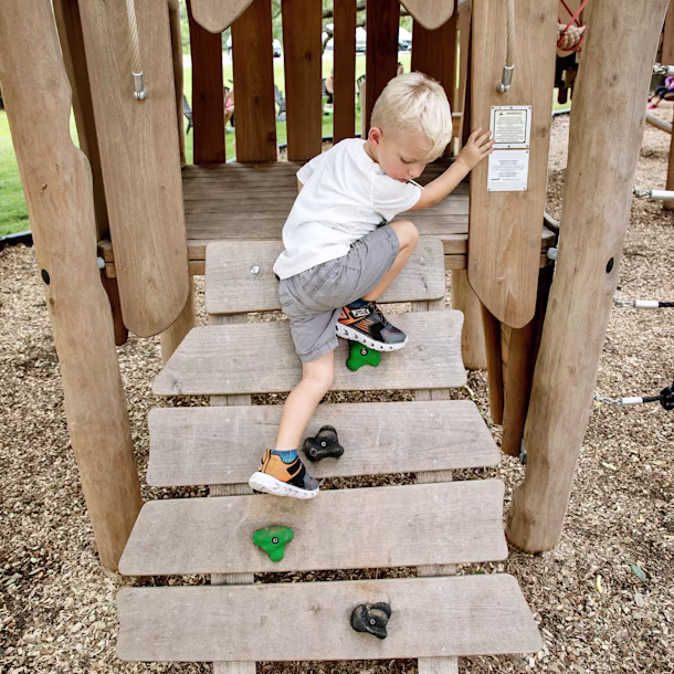 A young boy climbing on wooden play equipment. The floor below is natural coloured bark chippings. The aesthetic of your playground construction project can change the purpose and overall result. Be sure to choose wisely!