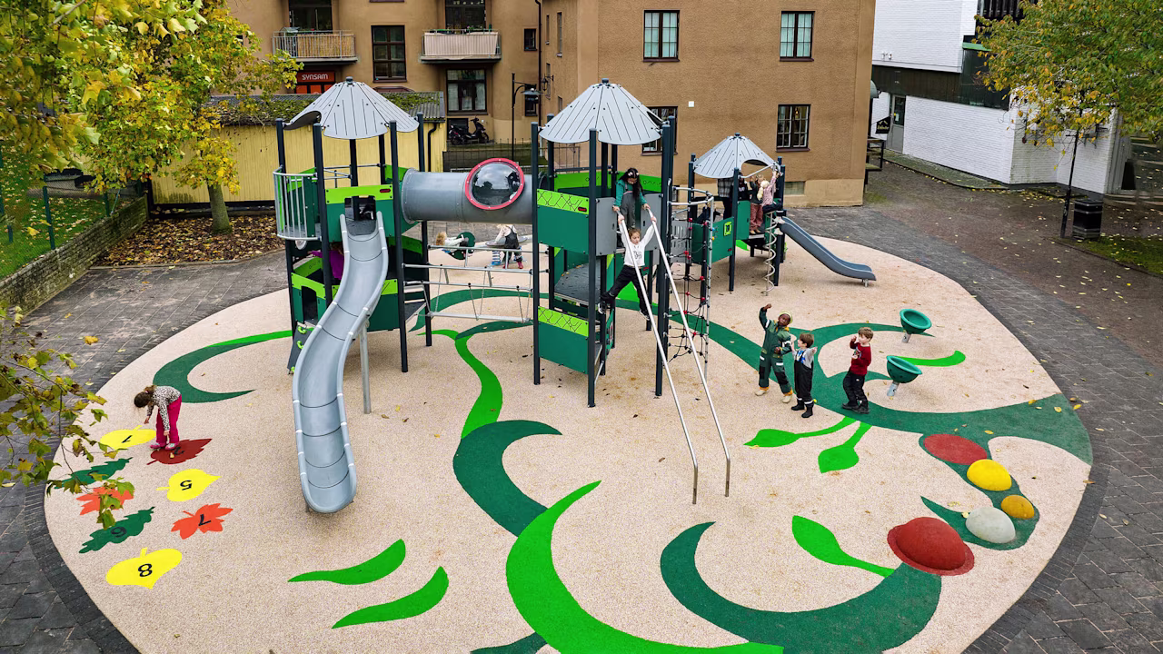 Children playing on a colourful, tree-themed playground with slides, climbing frames and numbered leaf markings in a city courtyard