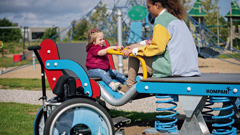 Child and adult sit on a blue spring seesaw in an inclusive playground, with a wheelchair parked beside the equipment.