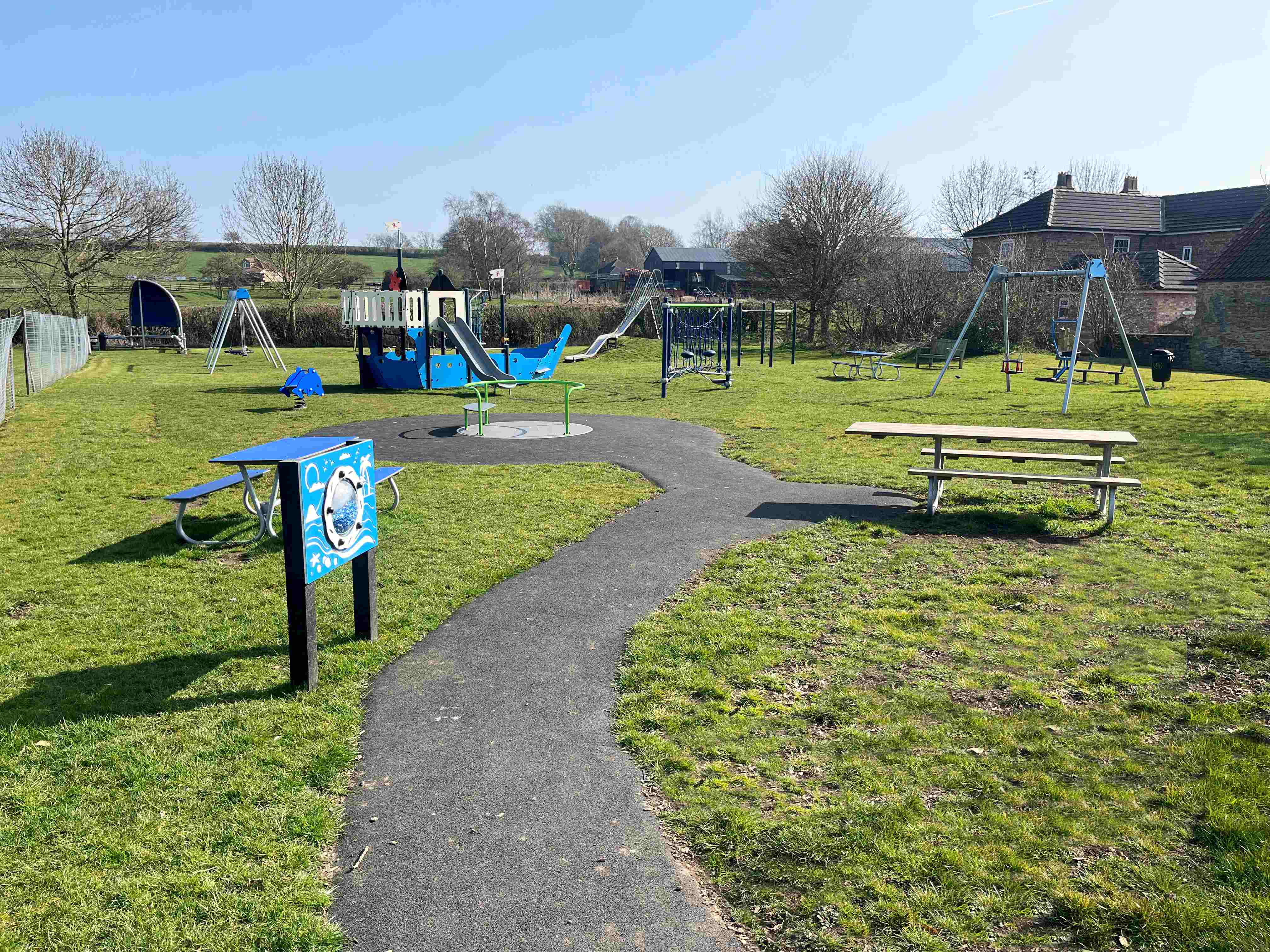 Sunny playground with blue ship-themed climbing frame, swings, picnic tables and a winding path on green grass near houses and fields
