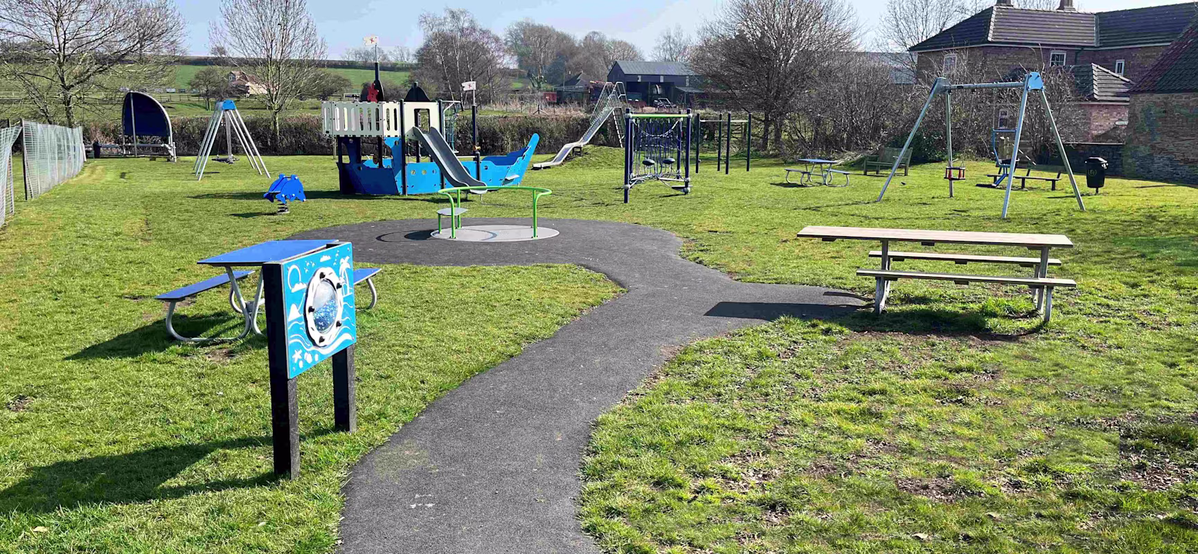 Sunny playground with blue ship-themed climbing frame, swings, picnic tables and a winding path on green grass near houses and fields