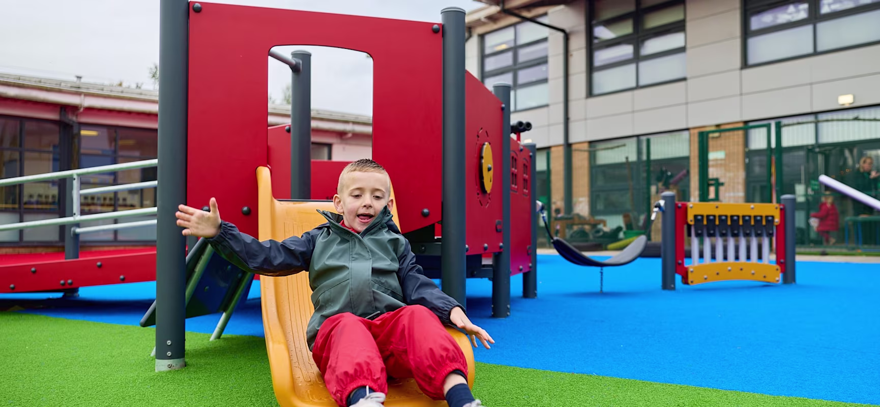 Child sliding down a yellow slide on a colourful playground with blue and green flooring outside a modern school building