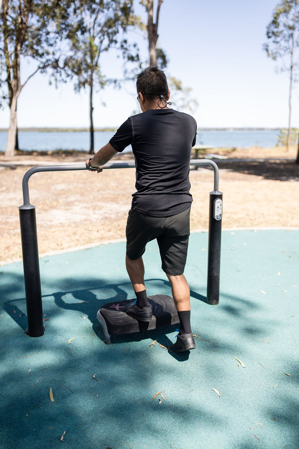 Man in black sportswear using an outdoor step exercise machine by a lakeside park, holding a metal rail under sunny trees