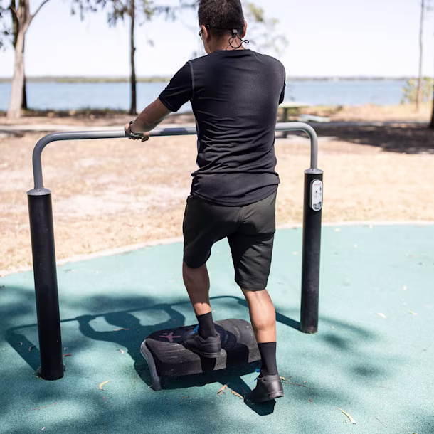 Man in black sportswear using an outdoor step exercise machine by a lakeside park, holding a metal rail under sunny trees