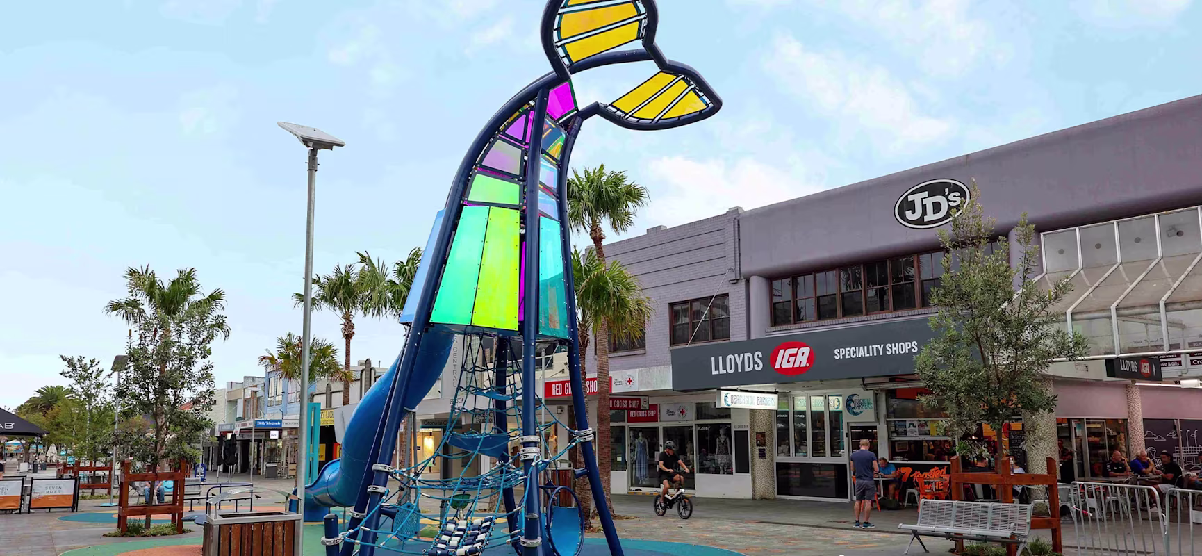 Colourful whale-tail themed playground structure in a town plaza, with shops, palm trees, a child on a bike and two seagulls nearby