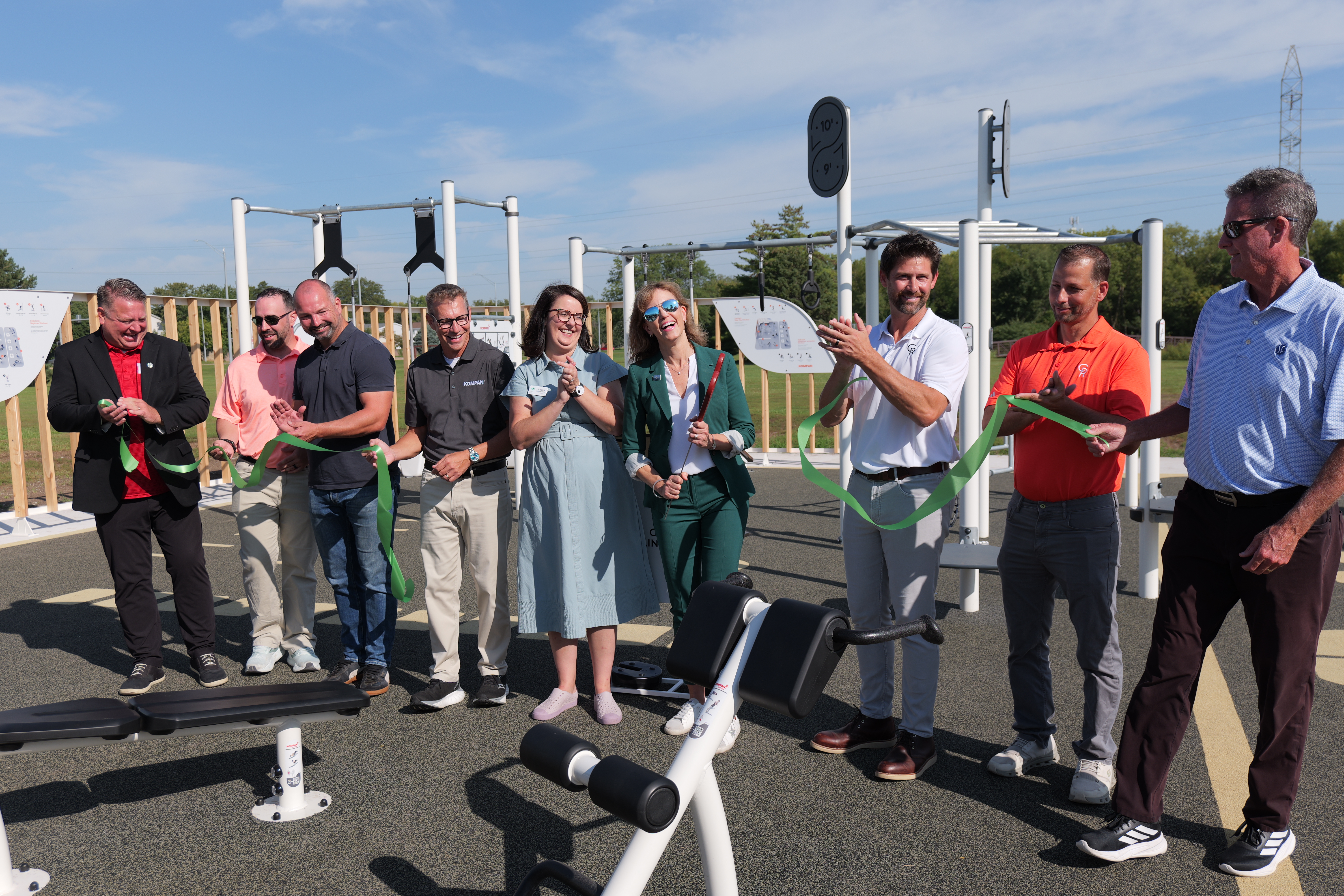 Group of people at an outdoor gym ribbon-cutting, holding a green ribbon among exercise machines under a clear blue sky.