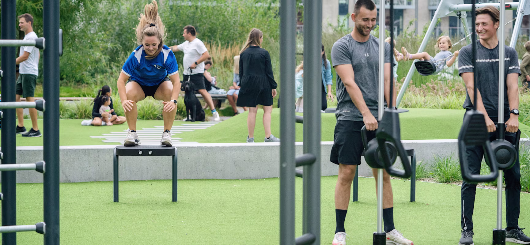 Woman using an outdoor leg press machine while a trainer kneels beside her, watching her form on a sunny sports ground