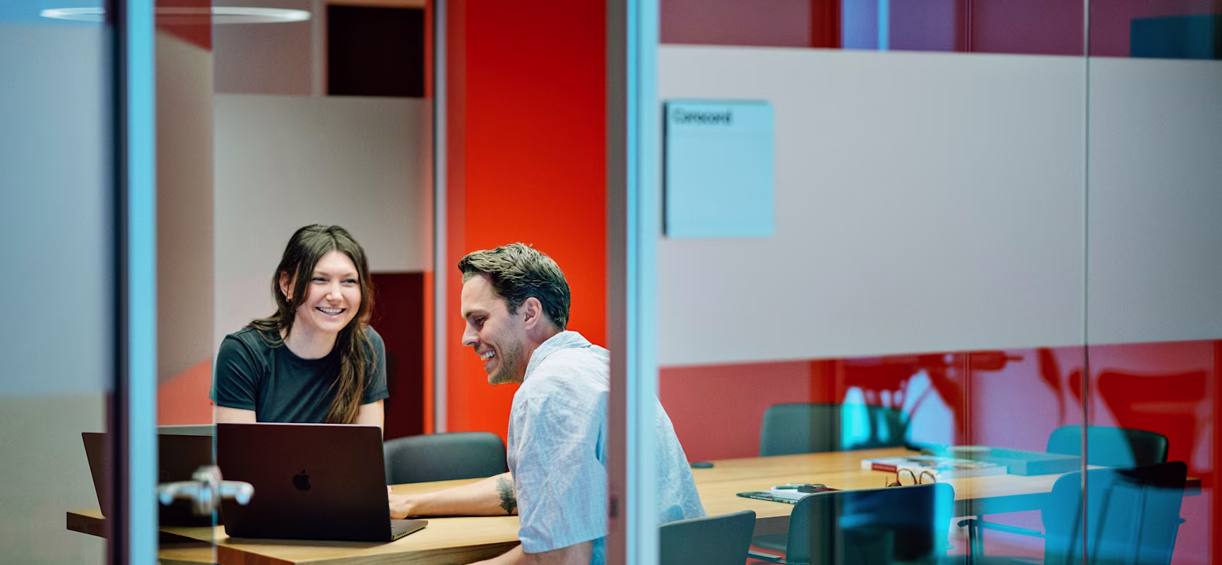 Two colleagues sit at a conference table with laptops in a glass-walled meeting room with red walls and circular ceiling lights