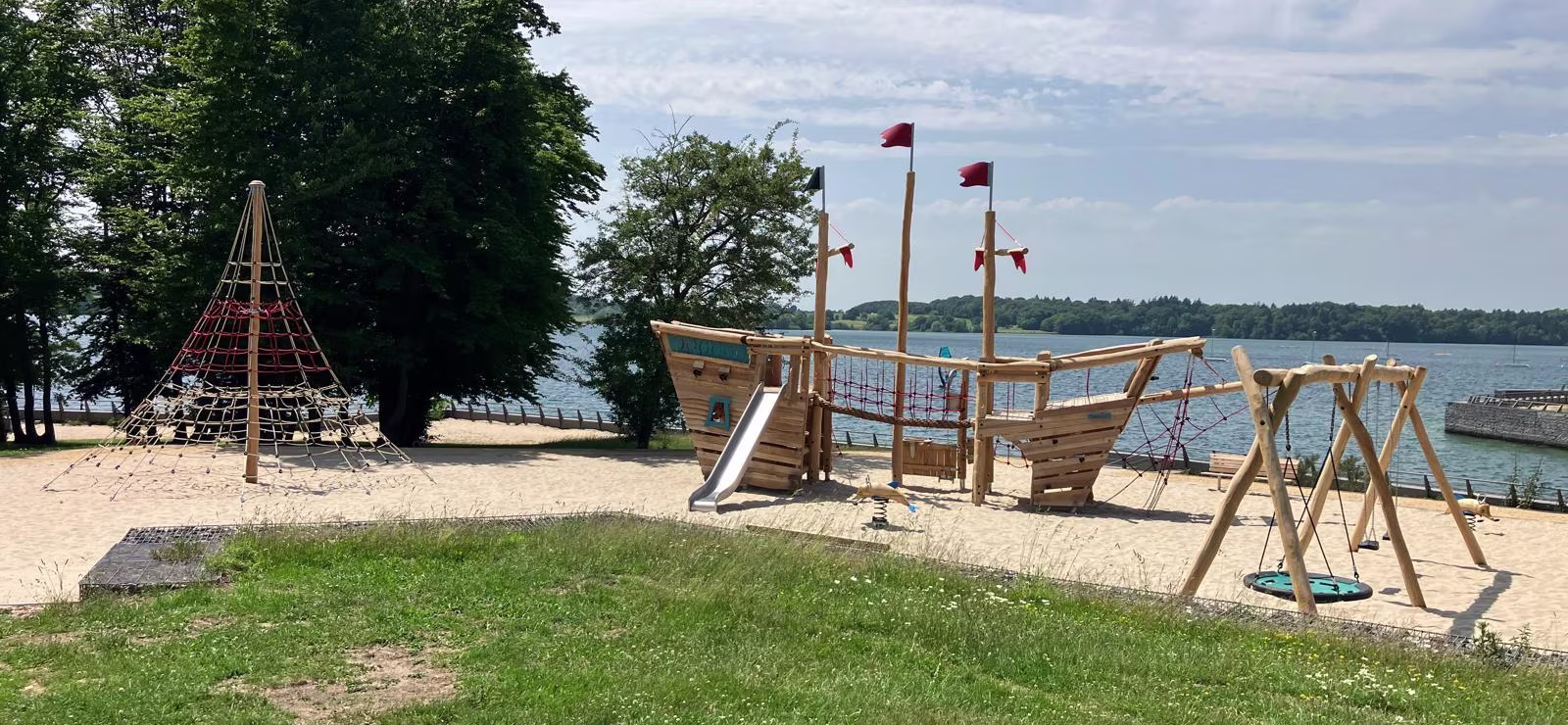 Sunny lakeside playground with a wooden pirate ship, swings, rope climbing net and trees on sandy shore by calm water