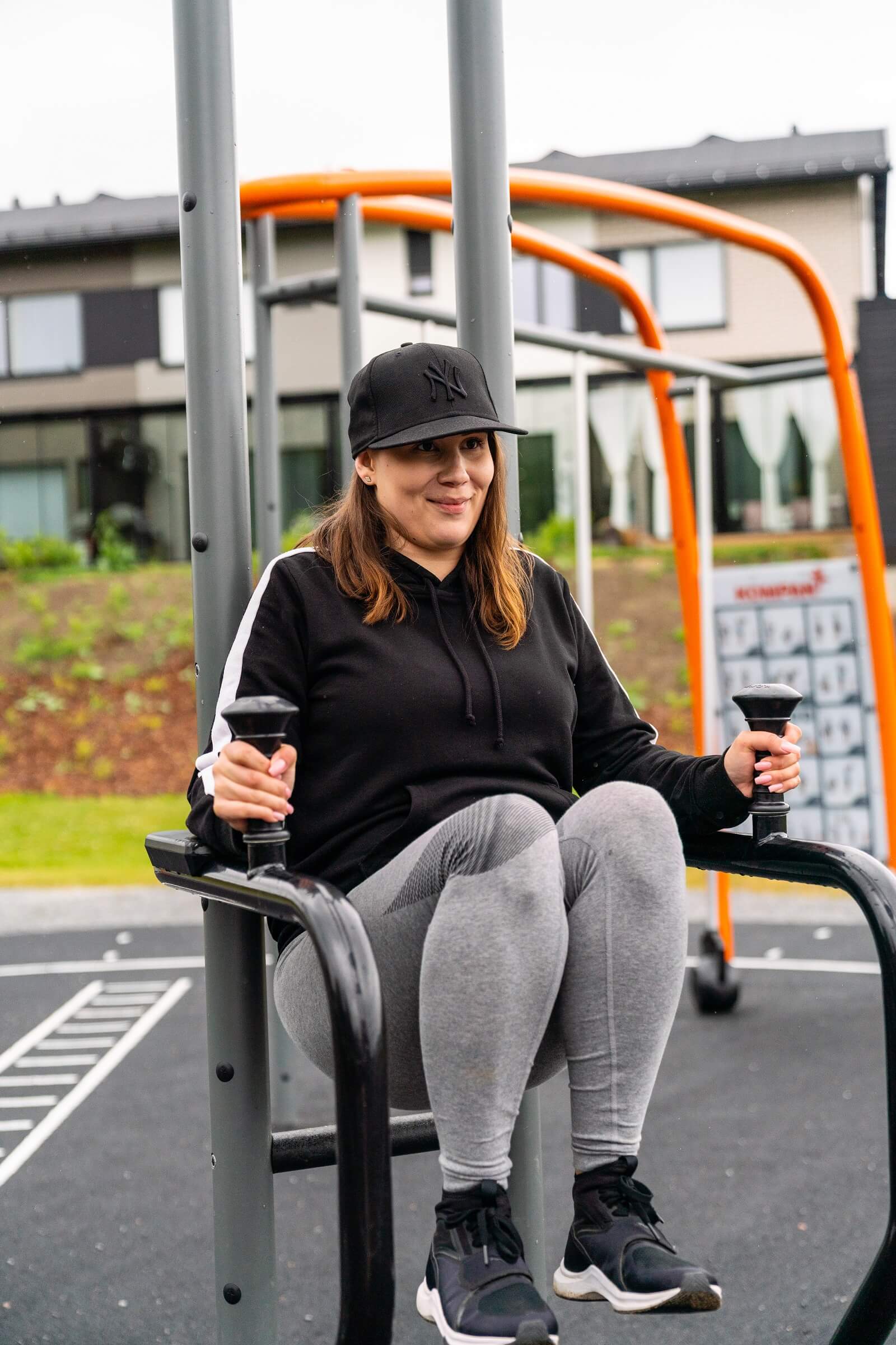 Person in black hoodie and cap exercising on outdoor gym equipment, lifting knees while gripping handles, with houses in background.