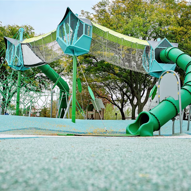 Modern playground with tall green climbing nets, elevated tunnels and twisting tube slides, set among trees on a rubberised surface