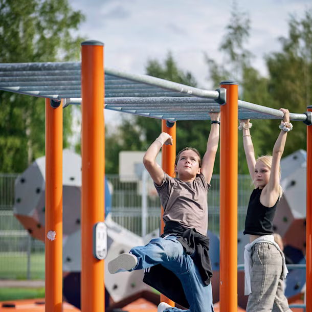 Children climb and hang from bright green playground bars in a school courtyard under a cloudy sky.