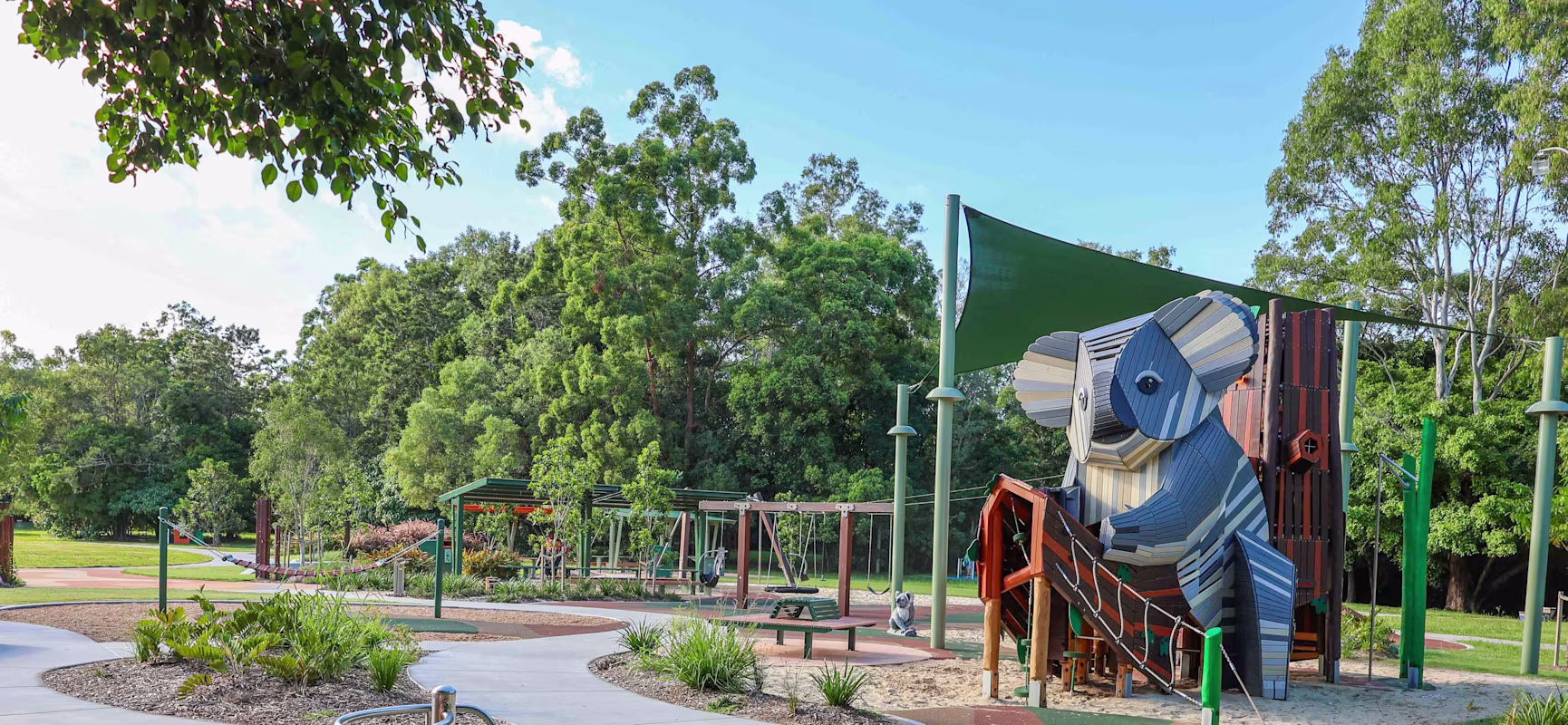Playground in a leafy park with a large blue koala-themed climbing structure, swings, paths and shade sails under a clear sky