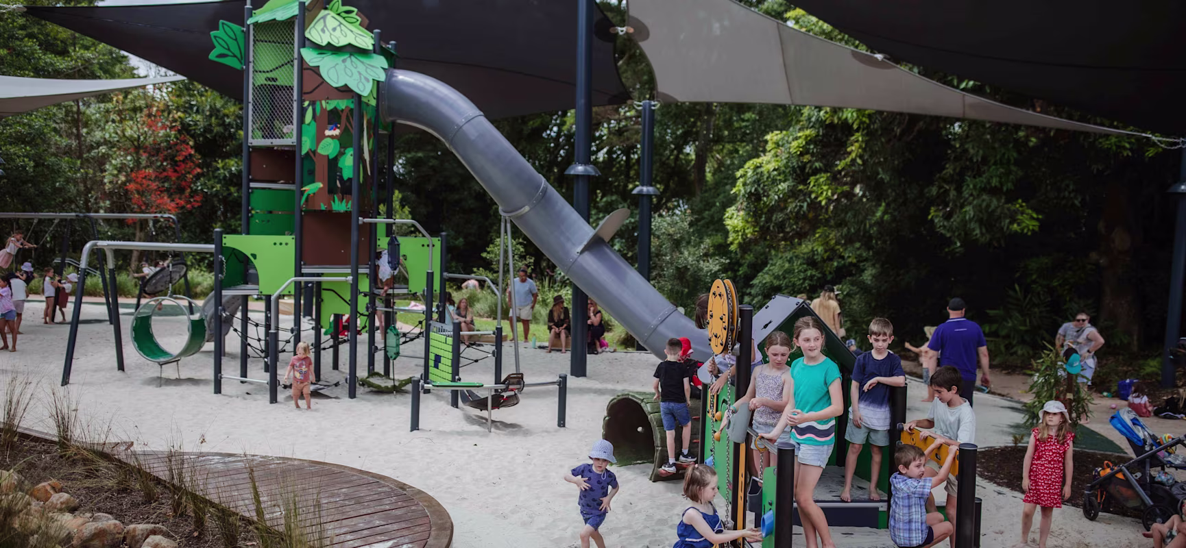 Children play on a shaded playground with tall slides and climbing structures, surrounded by sand, trees and supervising adults.