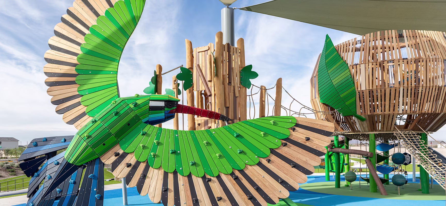 Colorful playground with a large wooden bird climbing structure beside treehouse towers and rope bridges under shade sails.