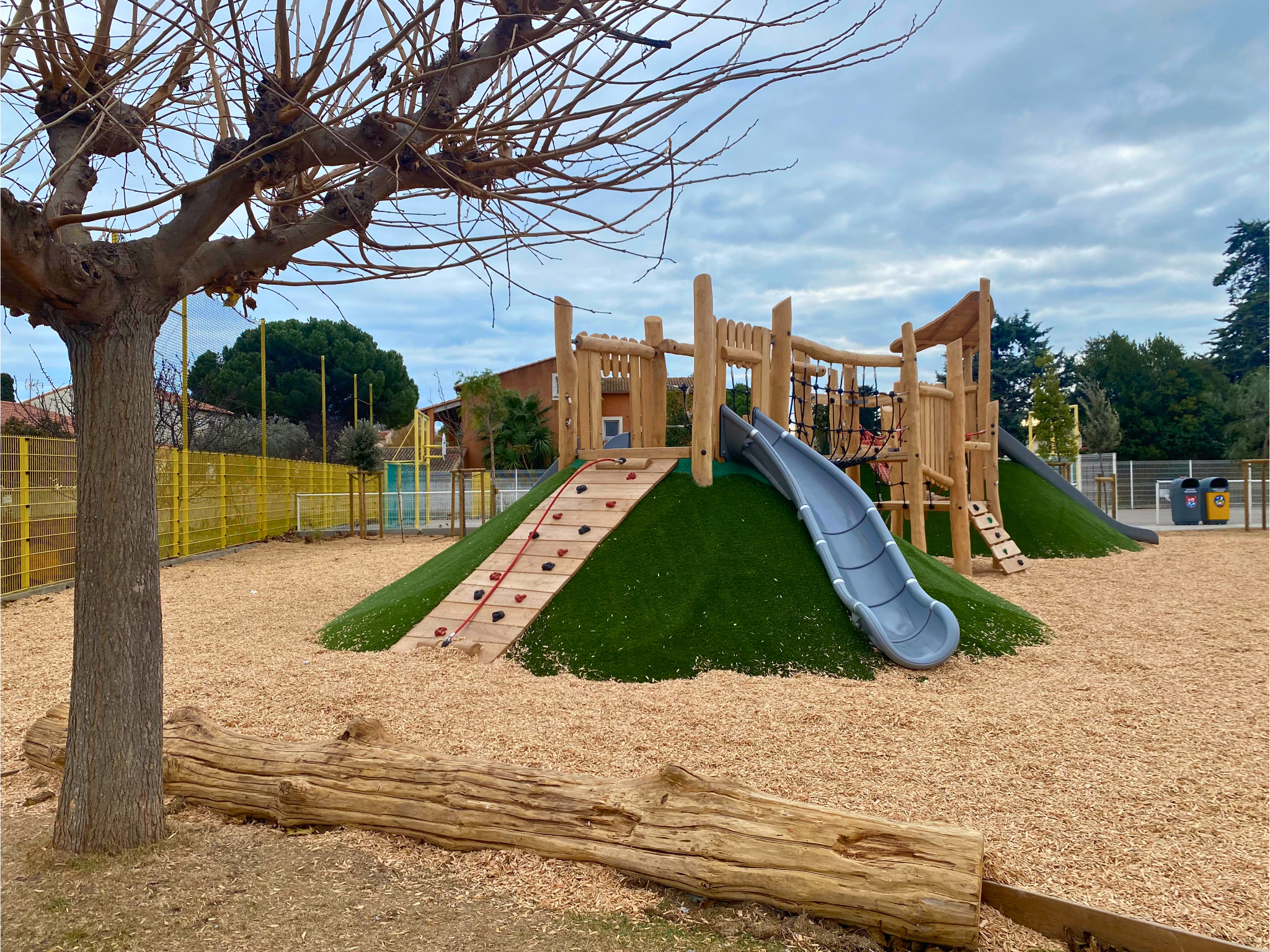 Outdoor playground with wooden climbing frames on grassy mounds, grey slides, woodchip ground, and bare trees under a cloudy sky