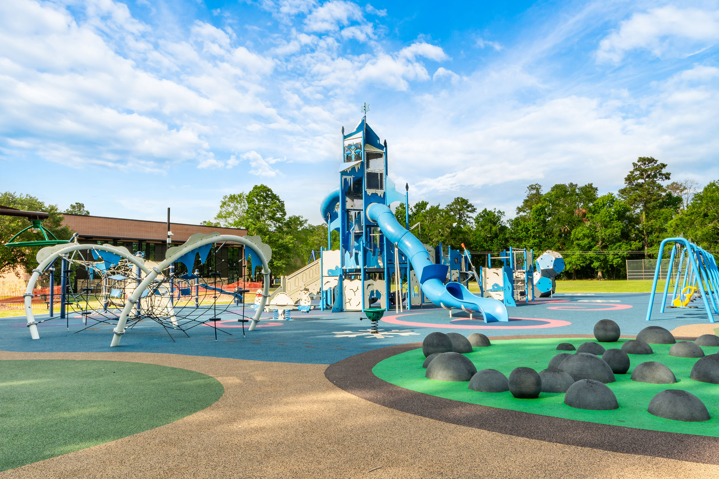 Colourful outdoor playground with tall blue slides, climbing nets and rubber mounds on open play surface under a bright sky.