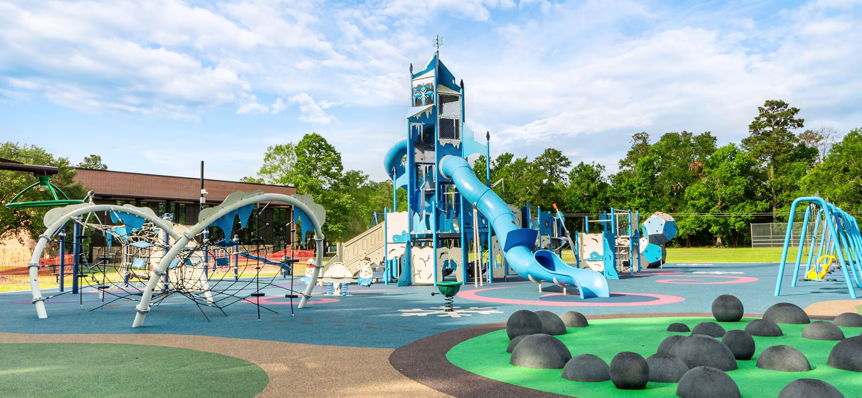 Colourful outdoor playground with tall blue slides, climbing nets and rubber mounds on open play surface under a bright sky.