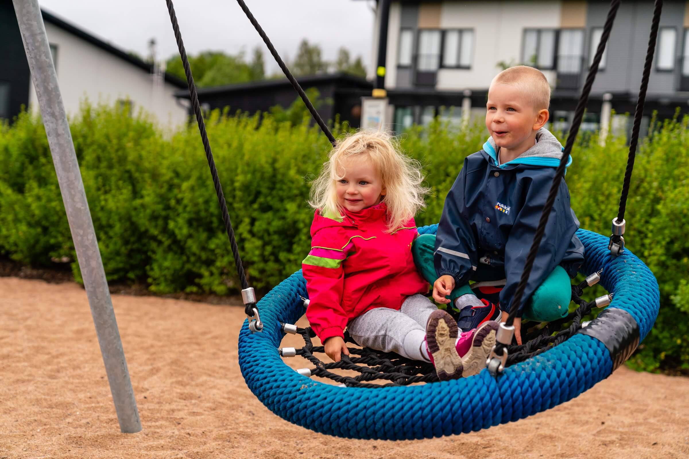 Two small children in colourful jackets sit together on a blue rope swing at a playground, with green bushes and houses behind them.