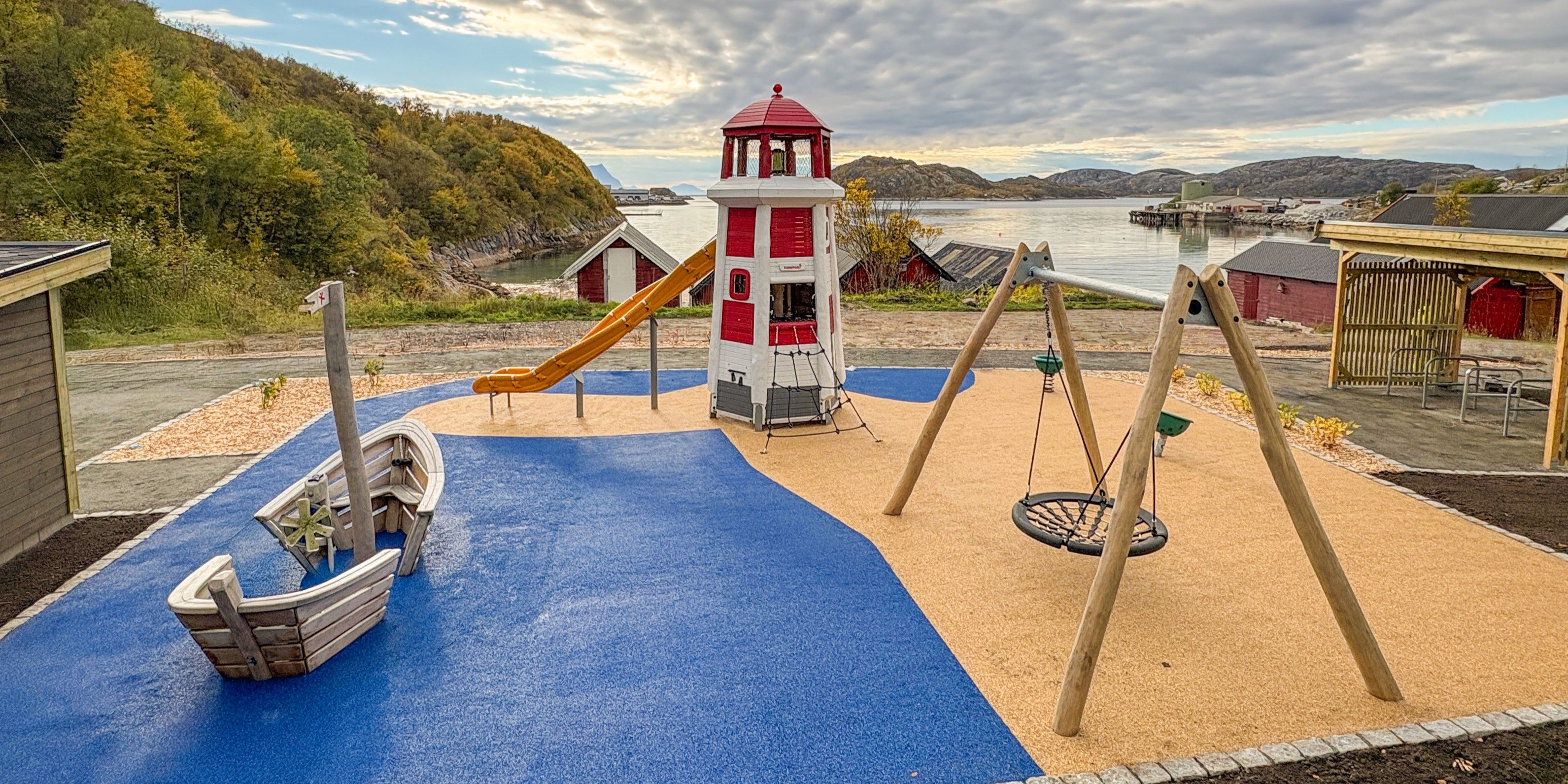 Coastal playground with a red-and-white lighthouse slide, wooden boat, and swings on blue and tan rubber near a calm fjord.