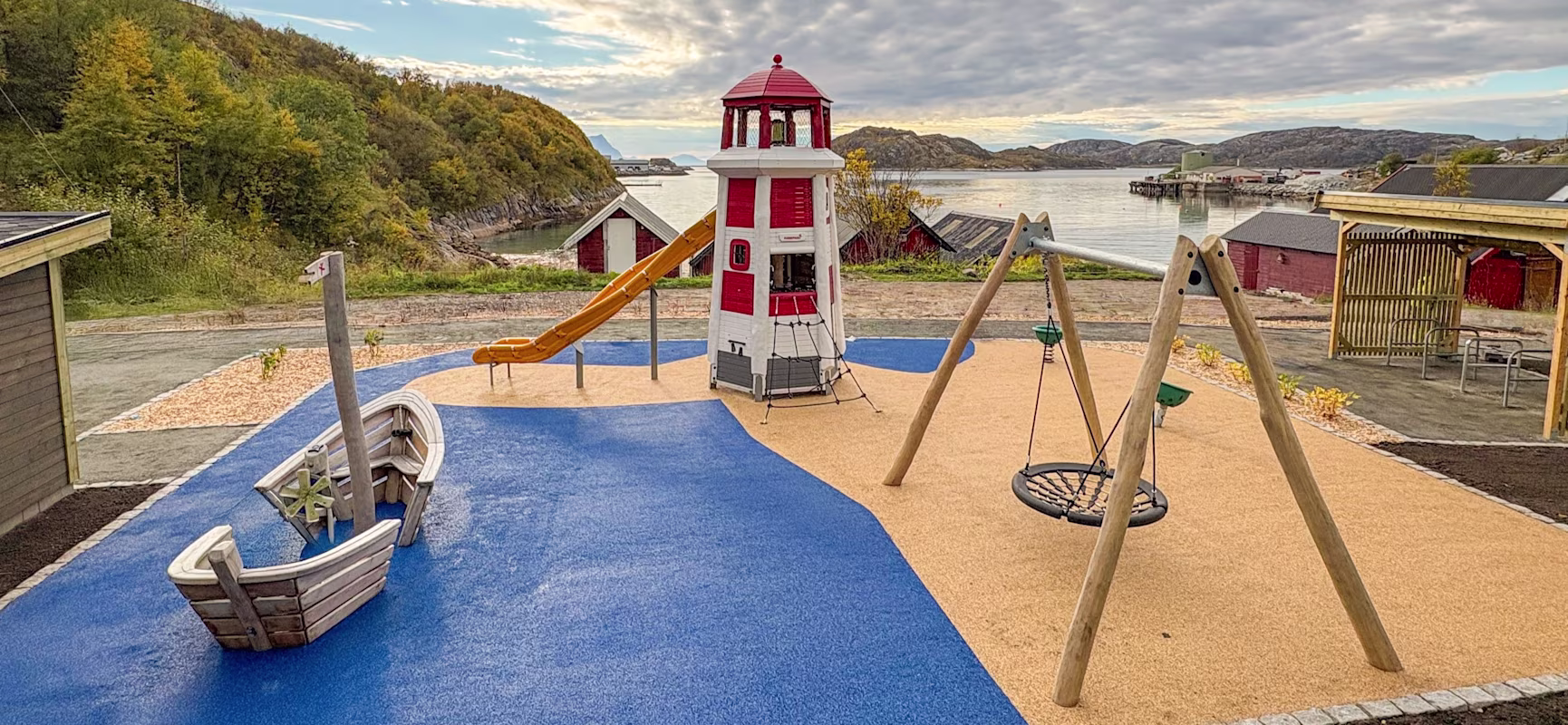 Coastal playground with a red-and-white lighthouse slide, wooden boat, and swings on blue and tan rubber near a calm fjord.