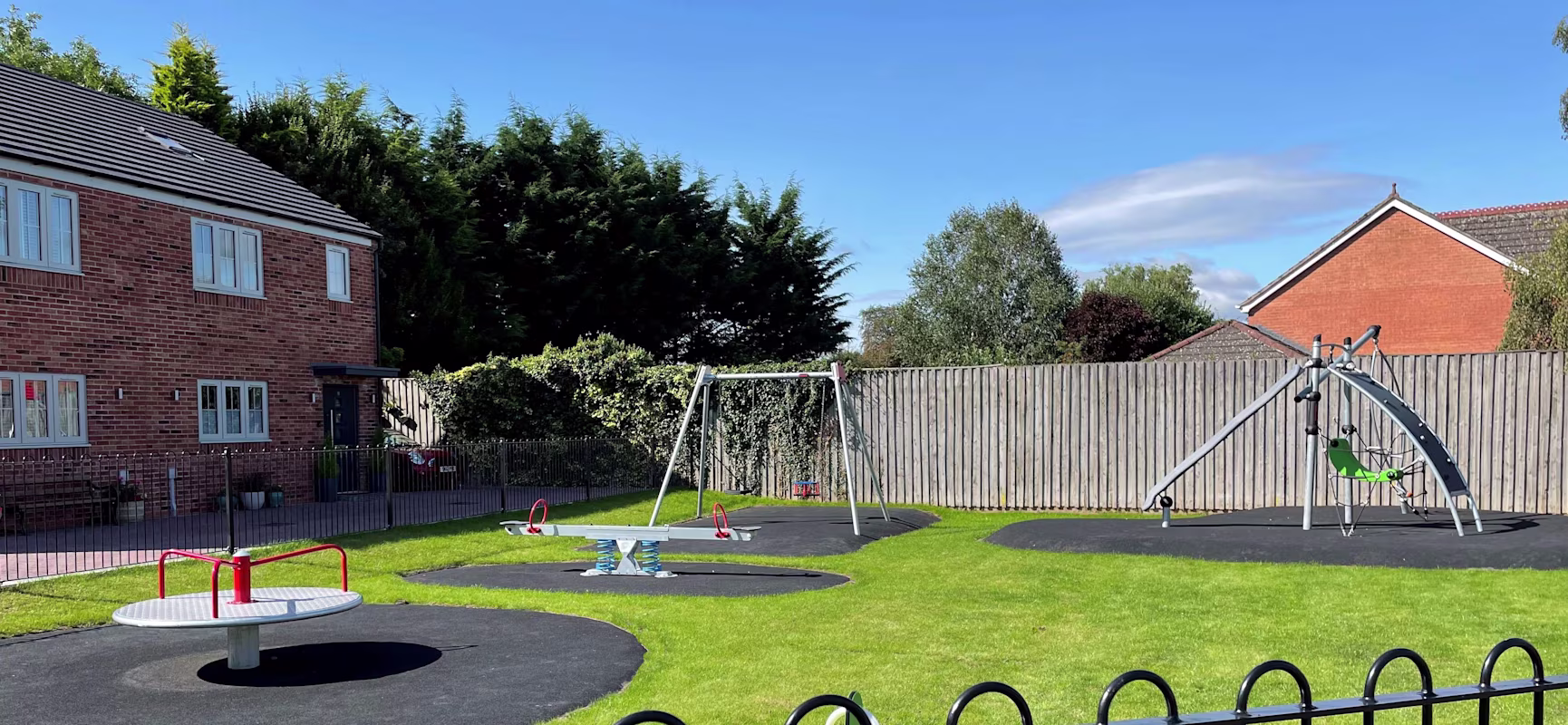 Small fenced playground with swings, roundabout, and climbing frame on grass, beside red-brick houses under a clear blue sky.