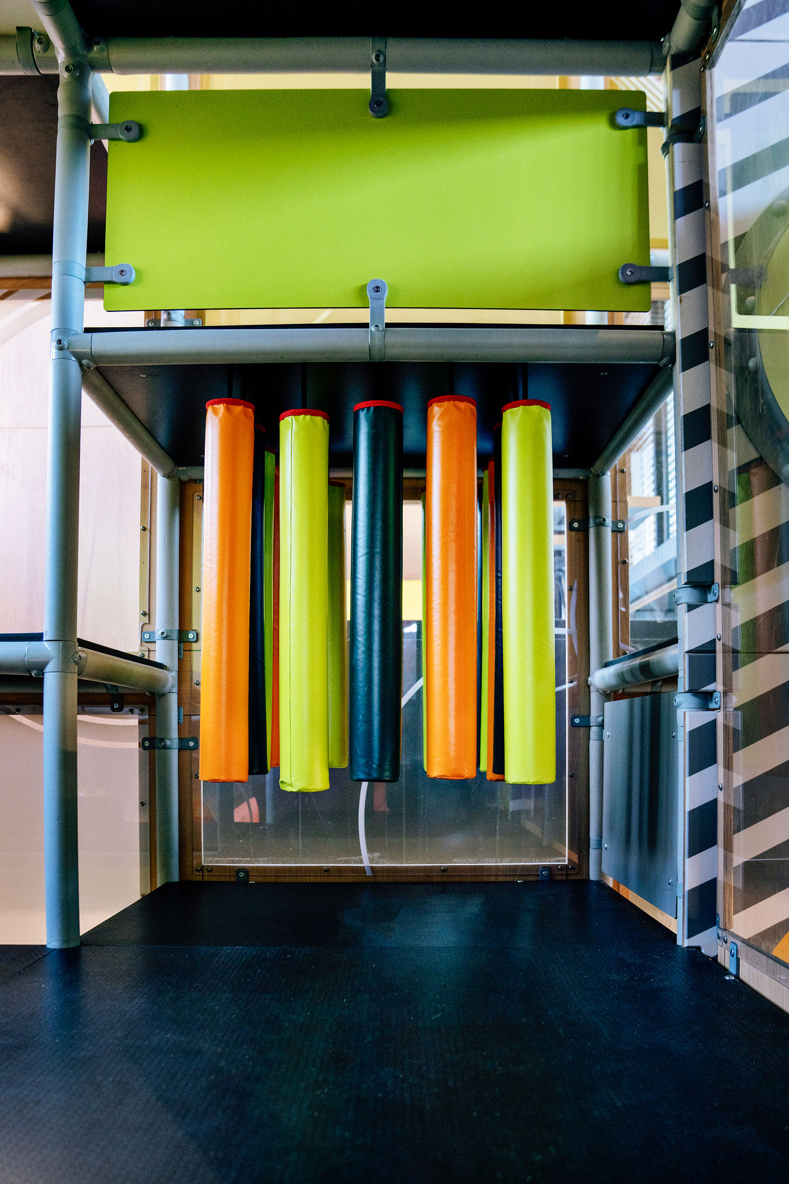 Indoor play structure with hanging padded tubes in orange, yellow and black, framed by metal bars and bright green panels