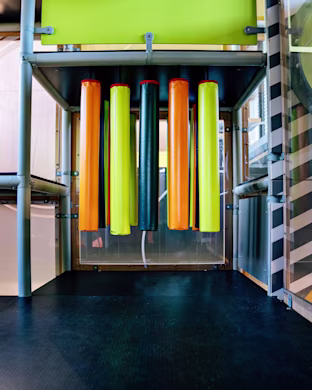Indoor play structure with hanging padded tubes in orange, yellow and black, framed by metal bars and bright green panels