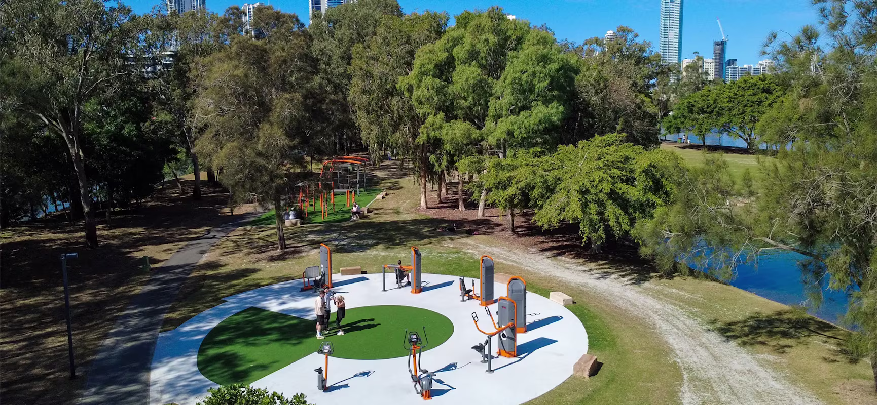 Outdoor gym with orange and grey equipment in a green park by a river, people exercising, tall city buildings in the background