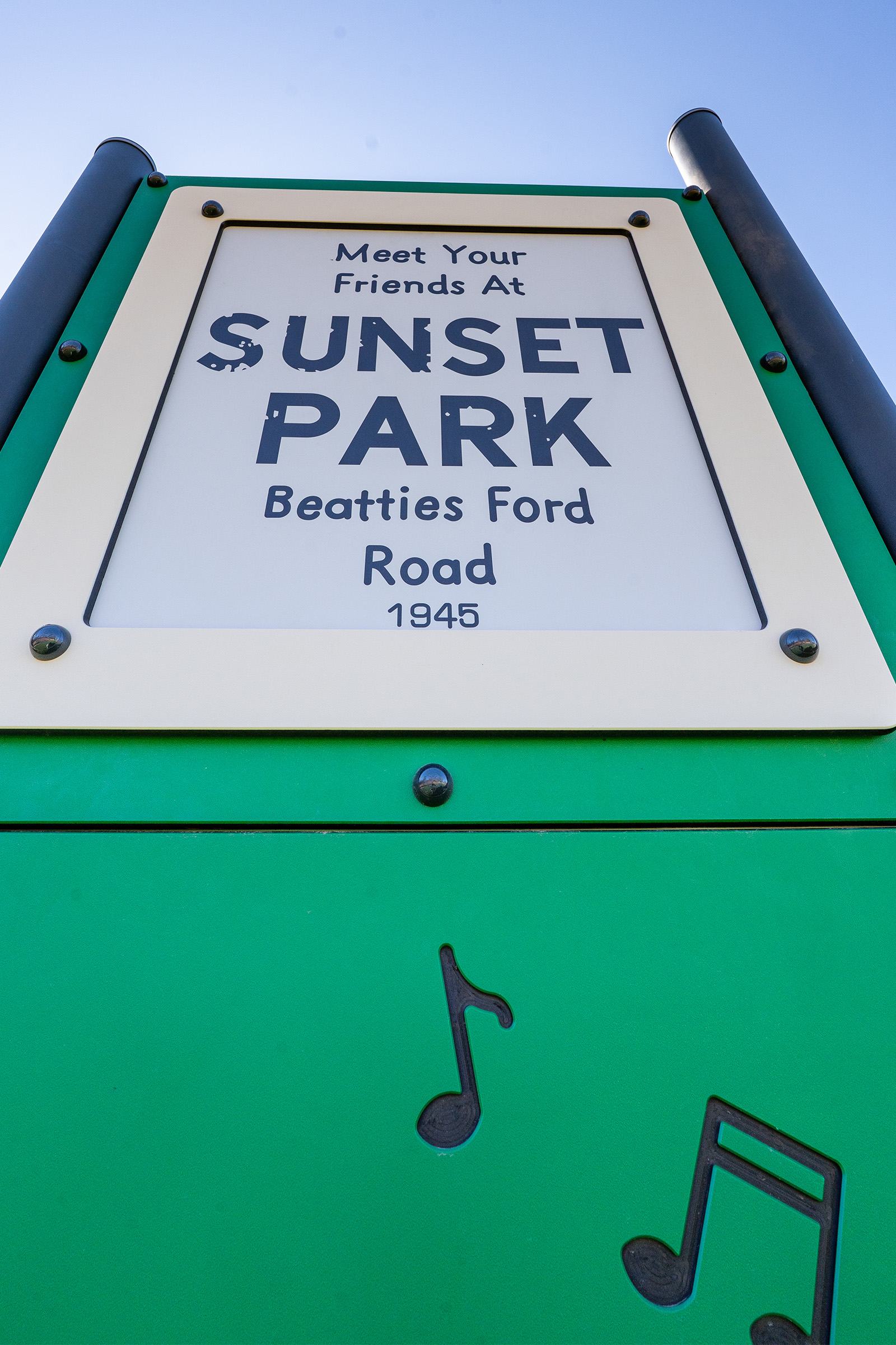 Close-up of green park sign reading “Meet Your Friends at Sunset Park, Beatties Ford Road, 1945,” with black musical notes at bottom.