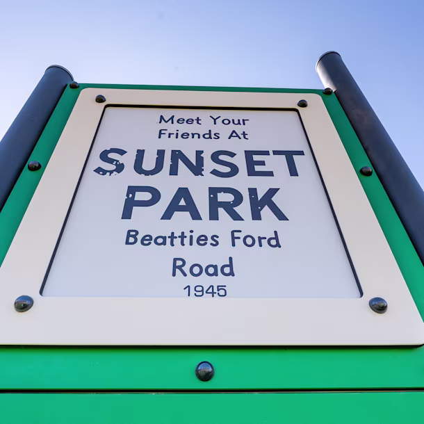 Close-up of green park sign reading “Meet Your Friends at Sunset Park, Beatties Ford Road, 1945,” with black musical notes at bottom.