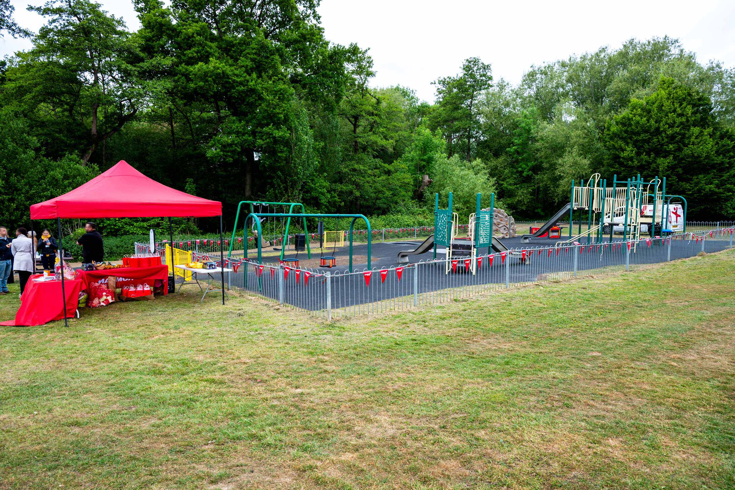 Playground with swings and climbing frames fenced with red bunting, next to a red event tent on grass, trees in background
