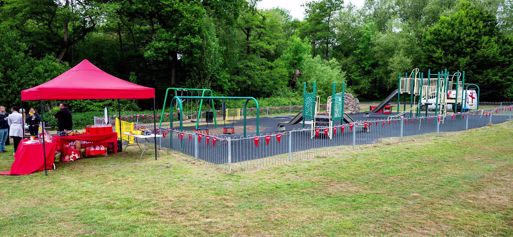 Playground with swings and climbing frames fenced with red bunting, next to a red event tent on grass, trees in background