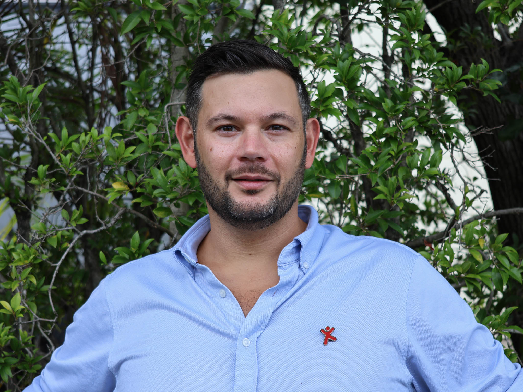 Person with blurred face in a light blue shirt stands outdoors in front of green leafy bushes.