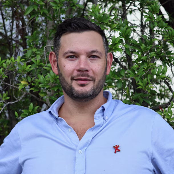 Person with blurred face in a light blue shirt stands outdoors in front of green leafy bushes.