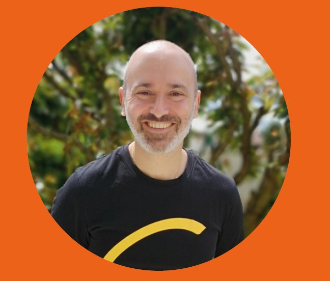 Bald person with blurred face in a black shirt with yellow curve, standing outdoors before green trees, framed by an orange circle.