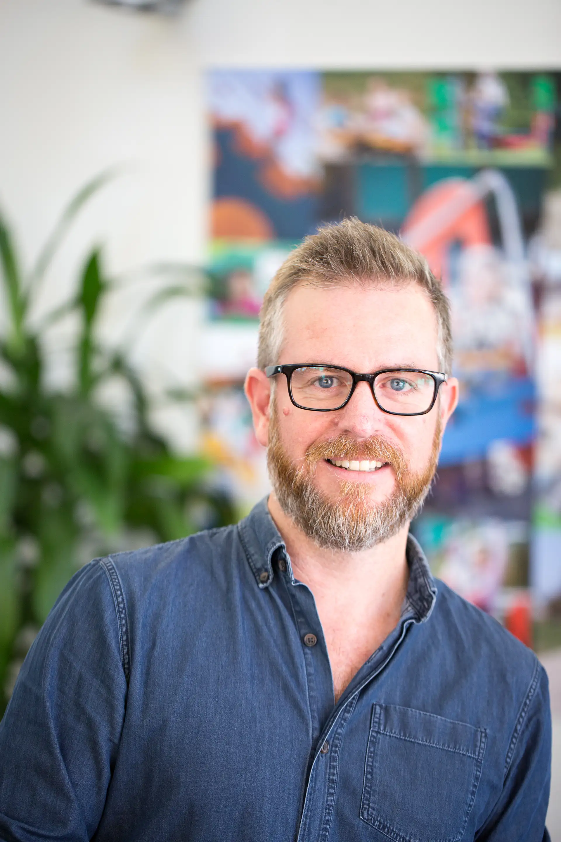 Person in a blue denim shirt with blurred face, standing indoors in front of colorful posters and green plants.