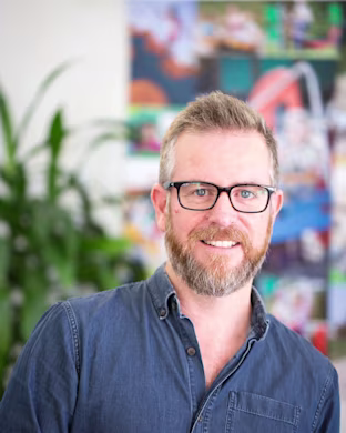 Person in a blue denim shirt with blurred face, standing indoors in front of colorful posters and green plants.