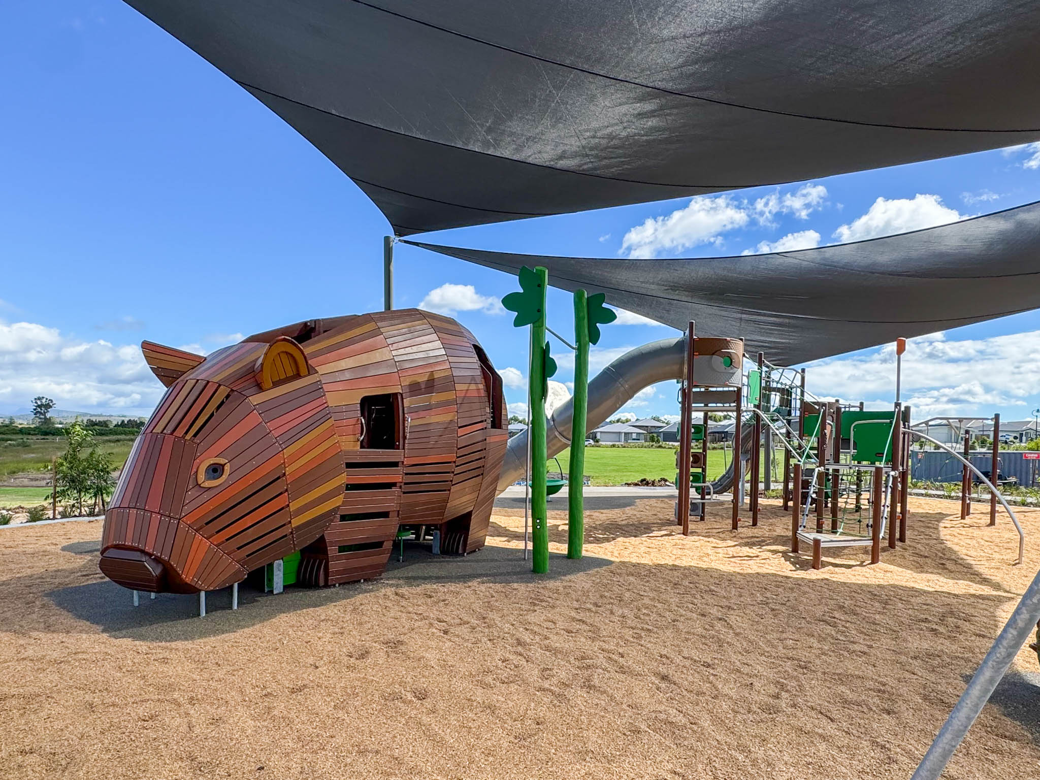 Shaded playground with a large wooden animal-shaped climbing structure and metal slides on tan mulch under a blue sky.