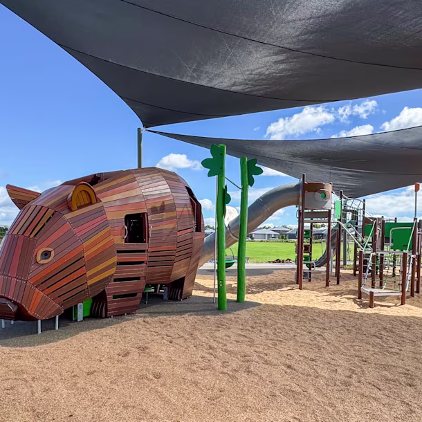 Shaded playground with a large wooden animal-shaped climbing structure and metal slides on tan mulch under a blue sky.