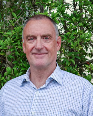 Person with blurred face in a checked shirt standing outdoors in front of leafy green bushes