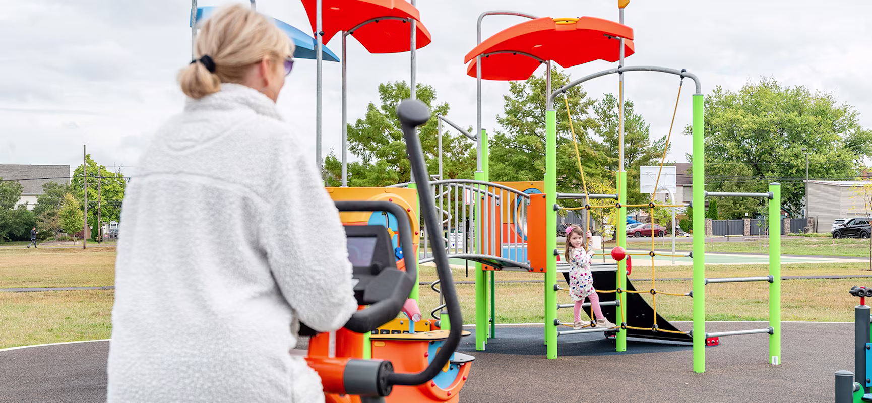 Woman exercising on a KOMPAN cross trainer while watching her daughter play on a KOMPAN playground.