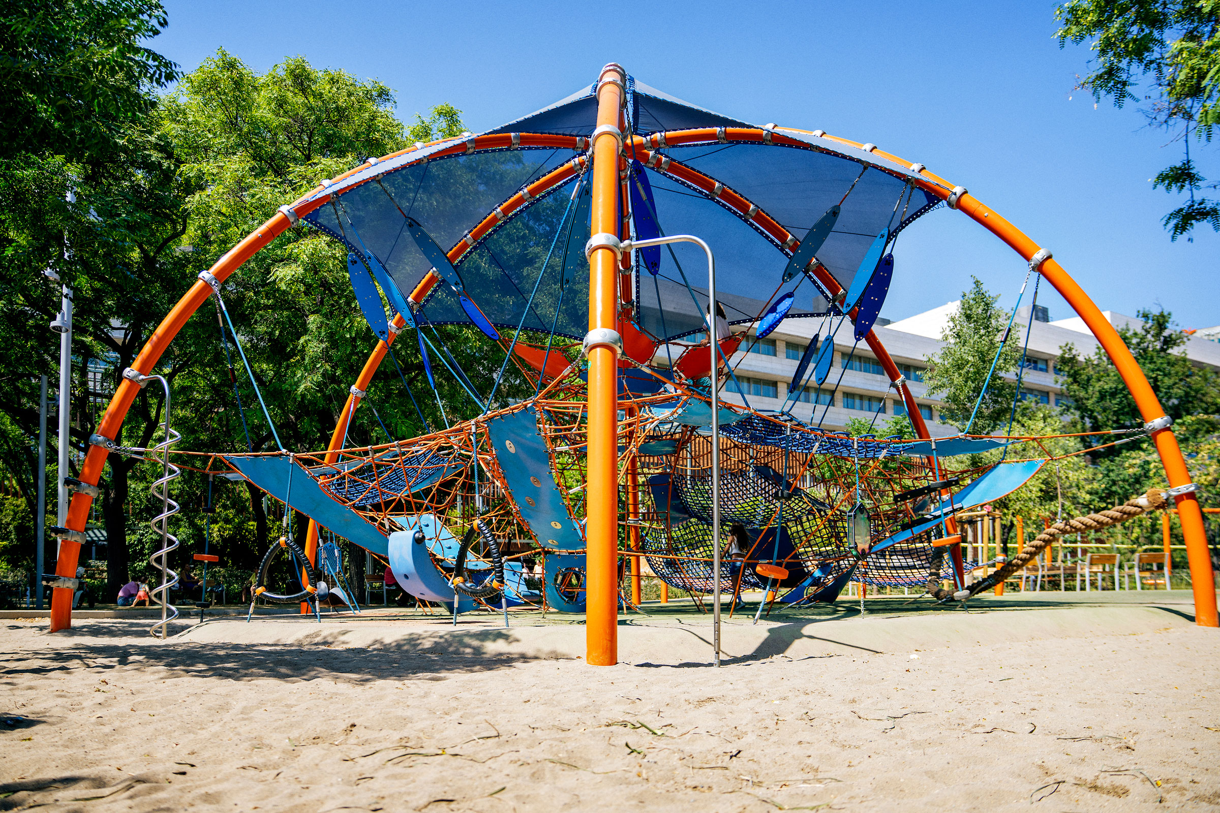 Modern playground with orange and blue climbing nets and slides on sand, surrounded by trees and buildings under a clear blue sky