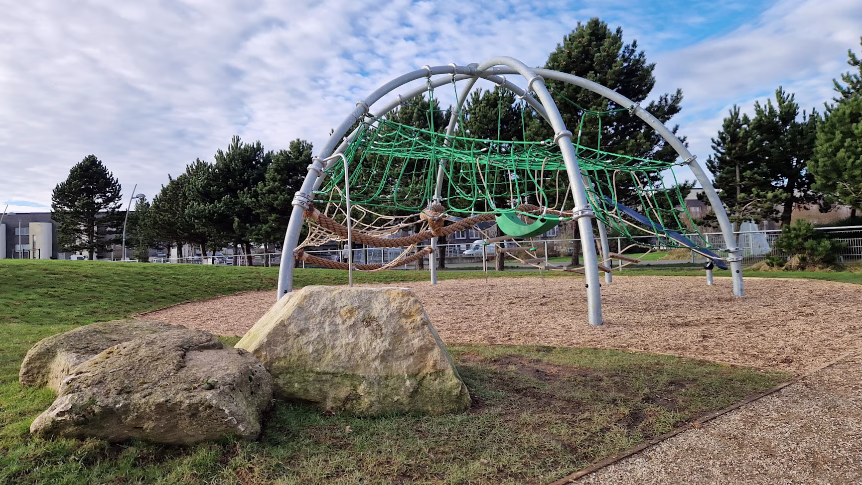 Modern playground with orange and blue climbing nets and slides on sand, surrounded by trees and buildings under a clear blue sky