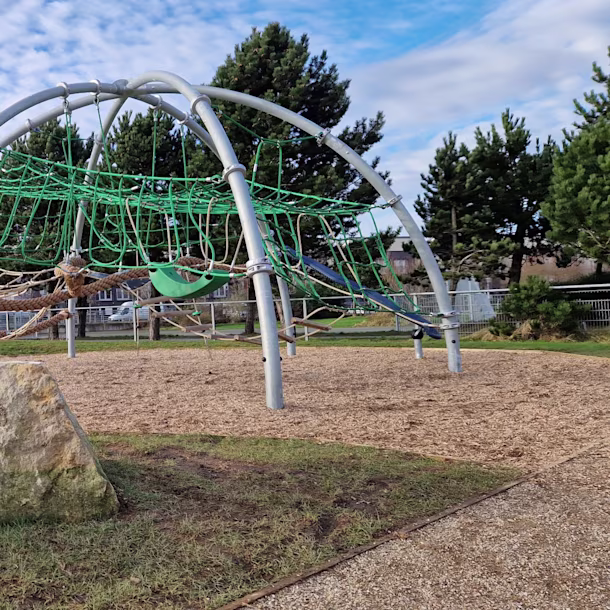 Modern playground with orange and blue climbing nets and slides on sand, surrounded by trees and buildings under a clear blue sky