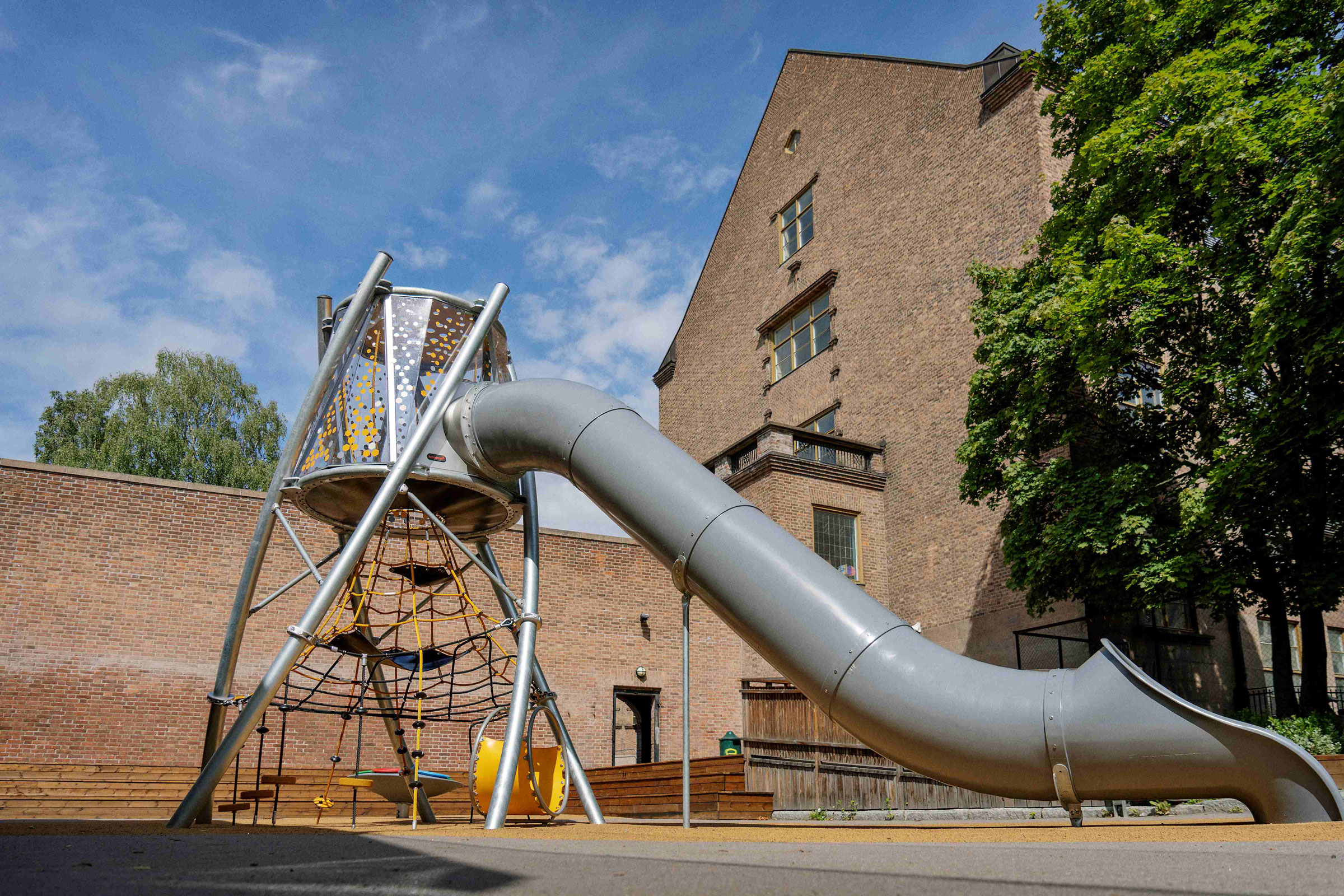 Modern playground with orange and blue climbing nets and slides on sand, surrounded by trees and buildings under a clear blue sky