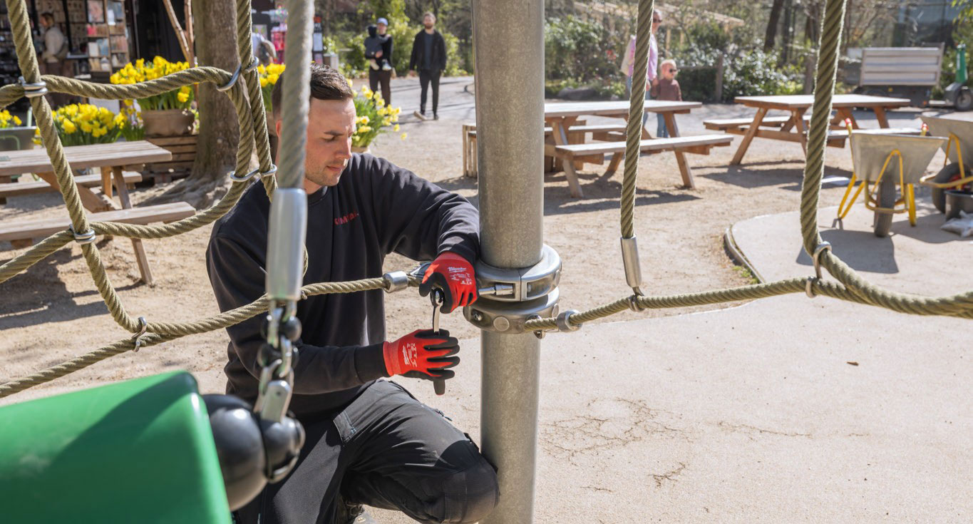 Worker in red gloves kneels by a metal pole, tightening rope netting at an outdoor playground with benches, flowers, and people in background