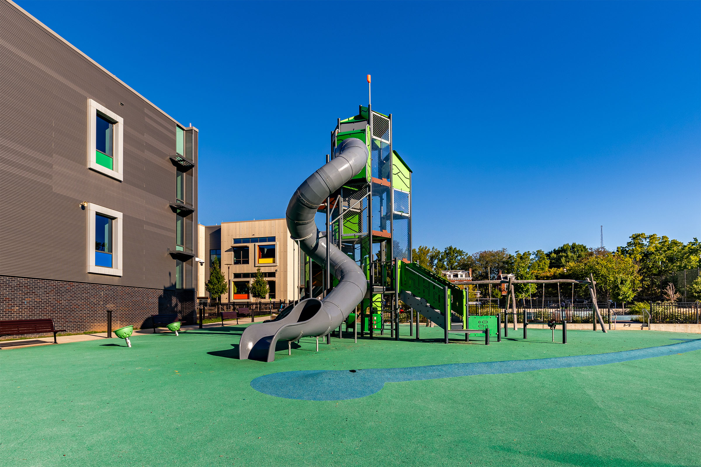 Empty modern playground with tall green climbing tower, spiral tube slide, swings and soft green surface beside colourful school buildings