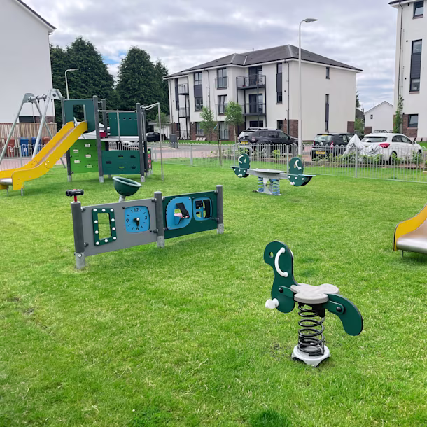 Modern fenced playground on green grass with slides, spring riders and play panels, set between parked cars and white apartment blocks