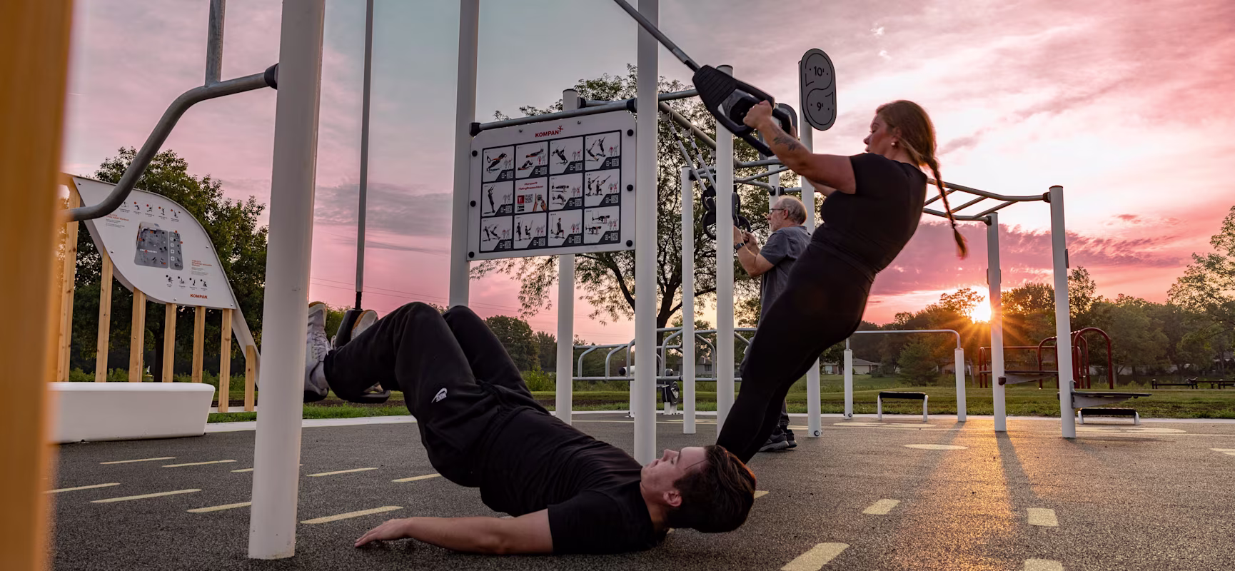 People exercise on outdoor gym equipment at sunset, one doing suspension rows, another performing floor leg curls under pink sky